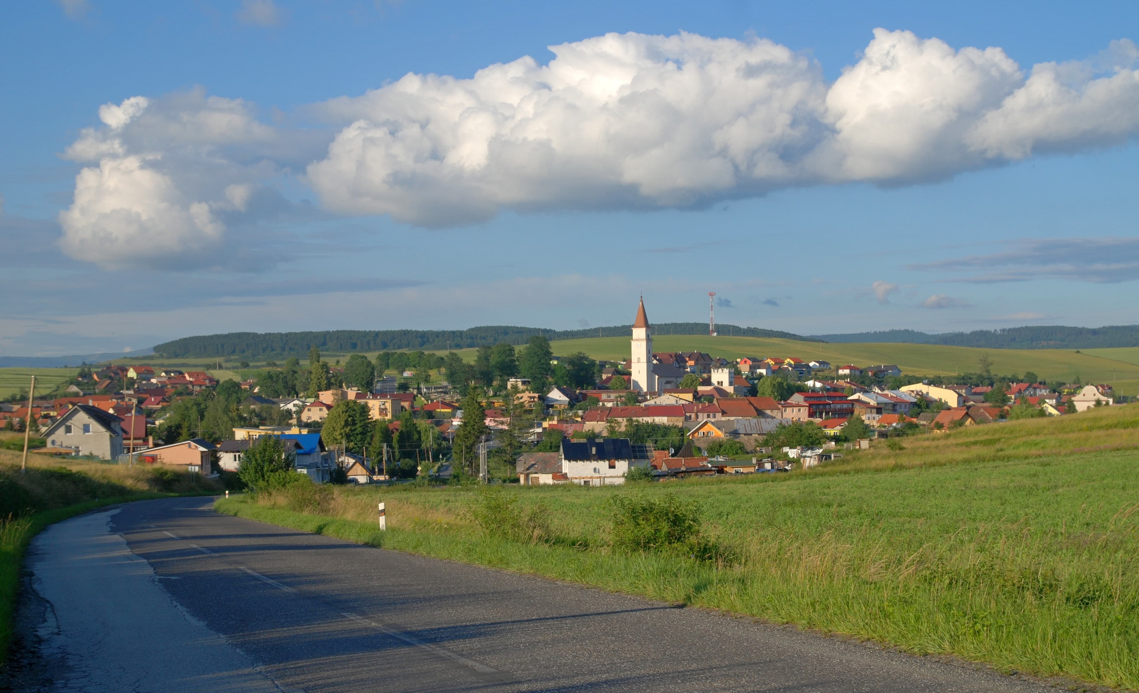 A scenic summer landscape featuring a winding road leading to a village with a prominent church tower, set against rolling green hills and a blue sky with cloud