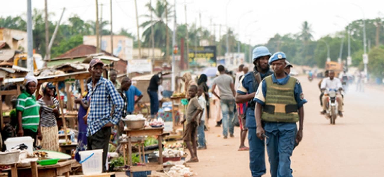 Street market scene with vendors, shoppers, and two uniformed officers walking on a dusty road