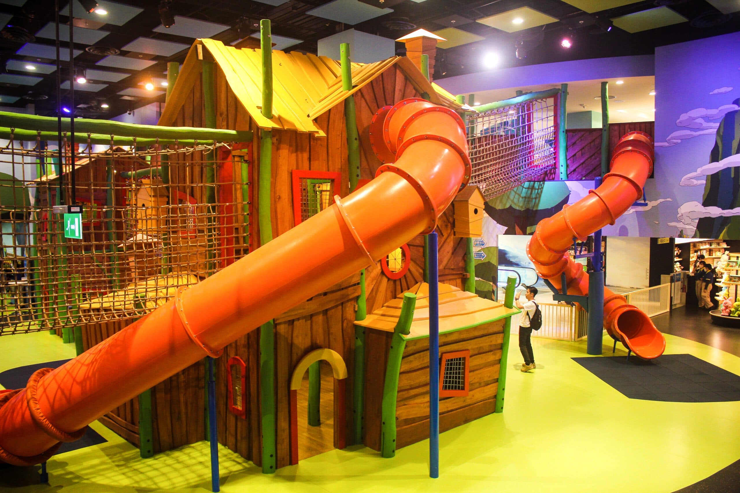 Colorful indoor children's play area featuring a large wooden structure with orange spiral slides, rope bridges, and a bright green floor.