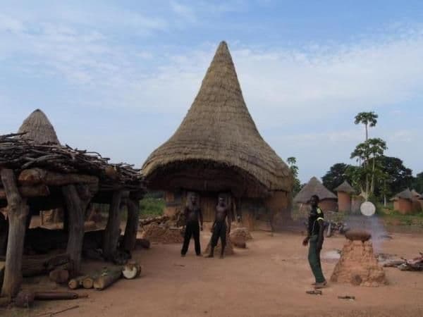 Wide shot of traditional thatched-roof huts in a dusty village setting with several people standing near structures and cooking fires