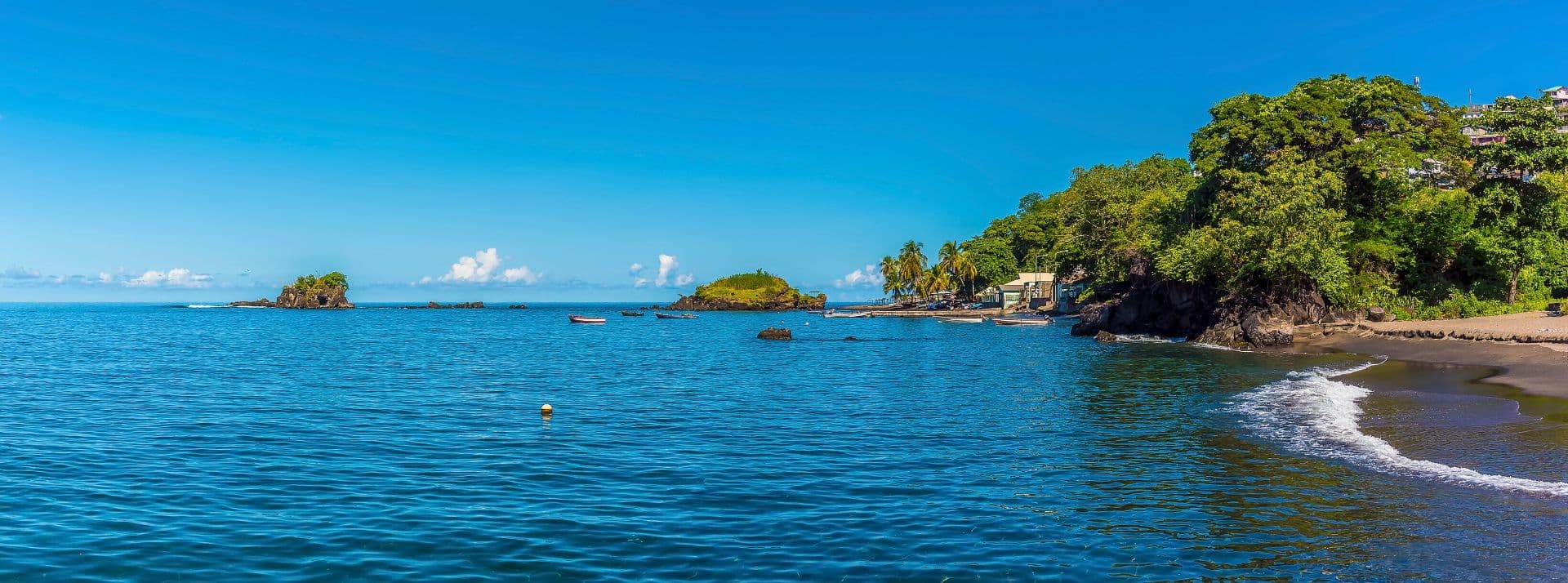 Calm blue bay water, small boats, rocky islets, a lush green headland, and a sandy shoreline near Richmond Beach, Barrouallie, Saint Vincent and the Grenadines.