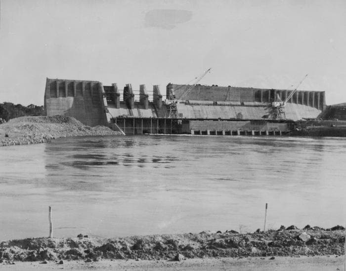 Black and white historical photograph showing the massive concrete structure of the Afobaka Dam under construction, with cranes visible against the sky.