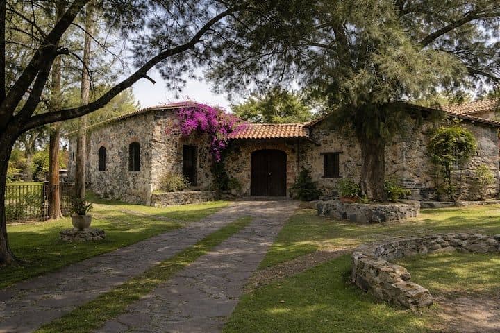 Stone rural house with terracotta roof, stone path, trees, and bougainvillea in a green highland setting.