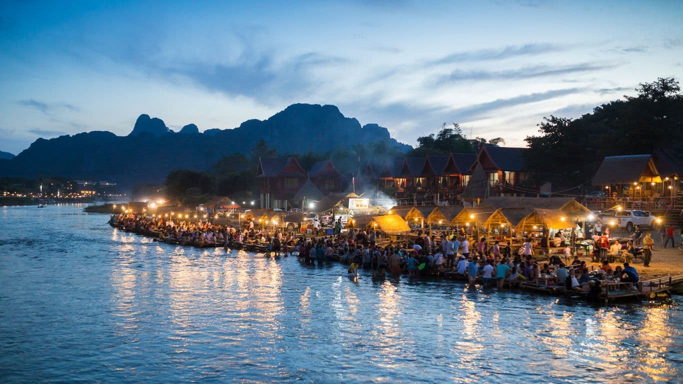Crowded floating market at dusk with thatched-roof restaurants and limestone karsts in the background.