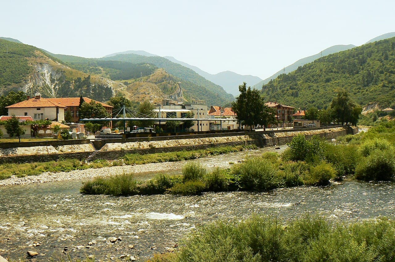 Wide exterior view of Krichim town featuring a shallow river, stone embankment, and buildings nestled against green mountains.