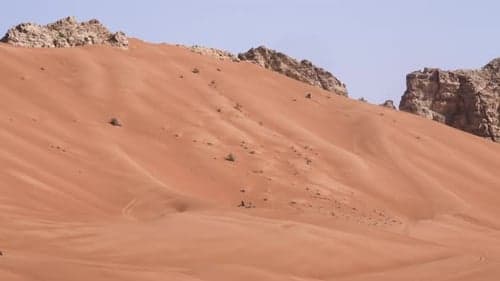 Wide shot of orange sand dunes with rocky outcrops under a clear sky, conveying heat and isolation in a desert landscape.