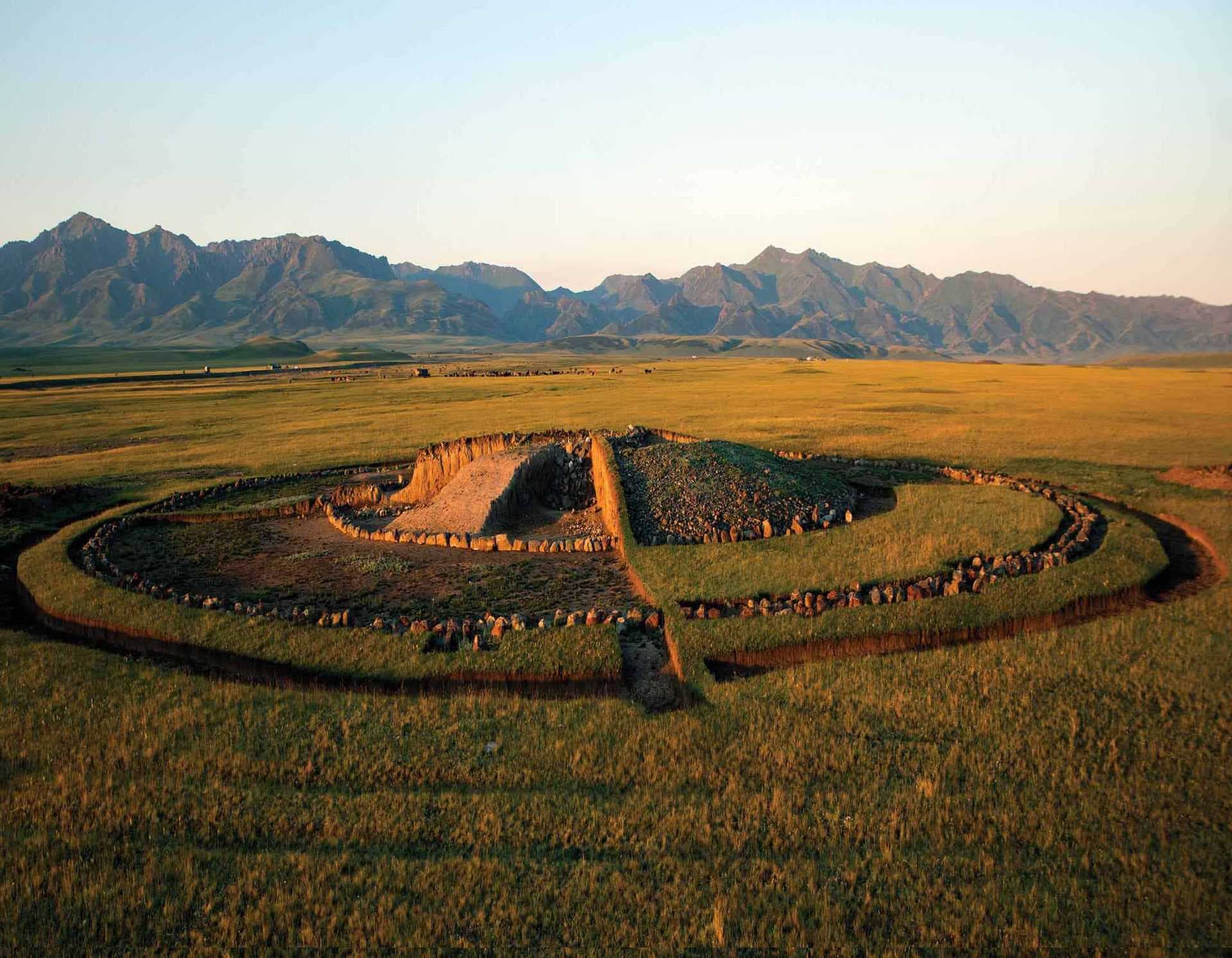 Aerial view of a grassy steppe with a circular stone structure, distant mountains, and warm golden light.