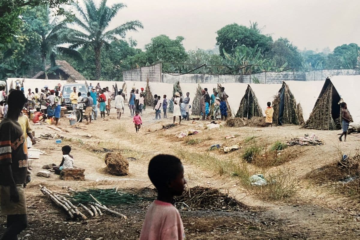 Rural village scene with people gathered near thatched structures and a white wall, set against lush tropical vegetation.