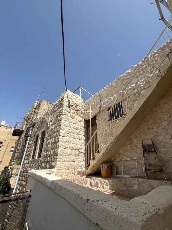 Exterior of an older stone guesthouse near Karak, Jordan, with arched windows, metal railings, and a staircase under a clear blue sky.