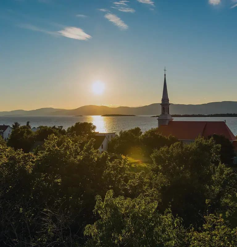 Sunset over water with a church steeple and red-roofed building in the foreground, framed by trees