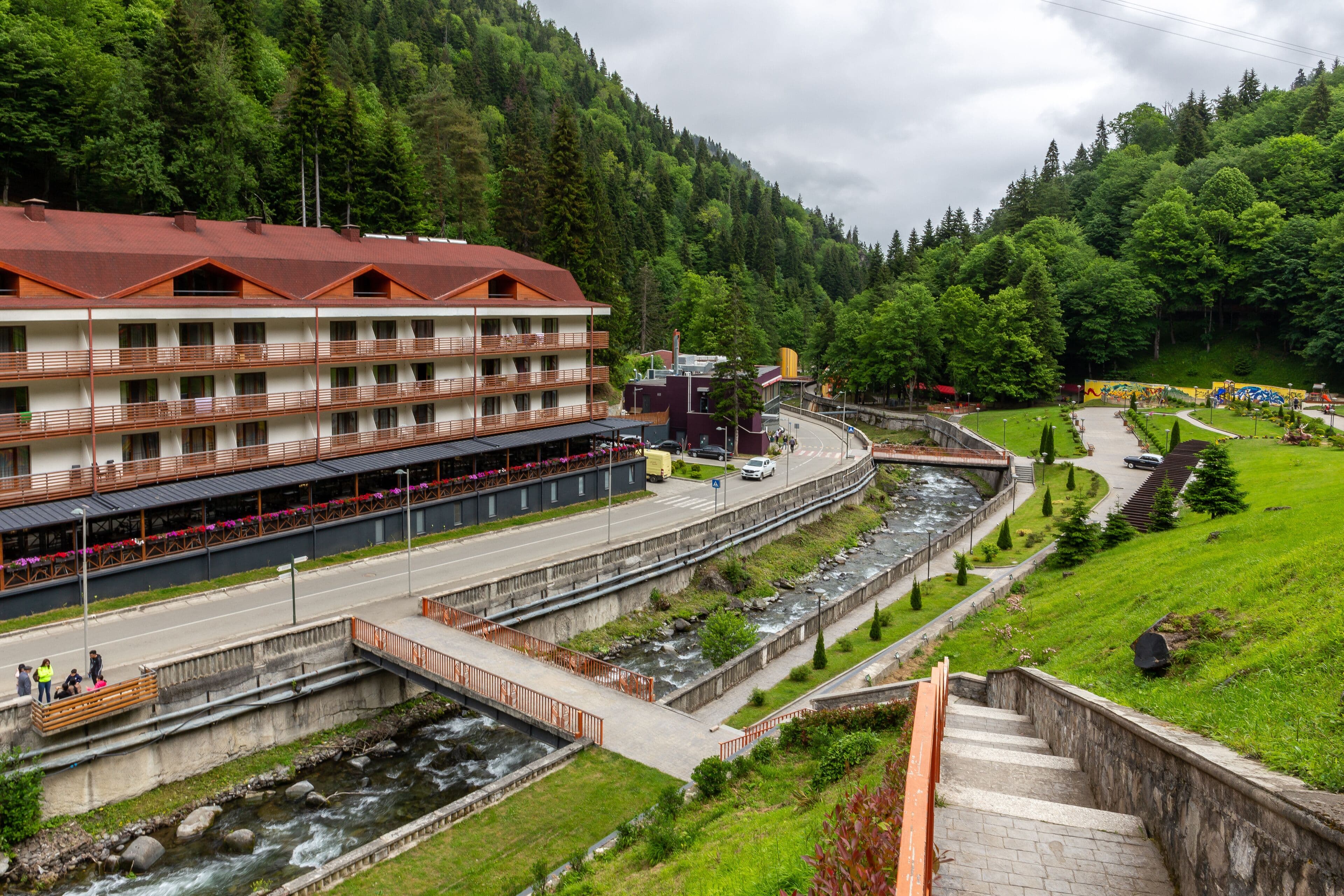 Wide scenic view of Baghdati municipality featuring a large hotel, a flowing river, and lush green forested hills.