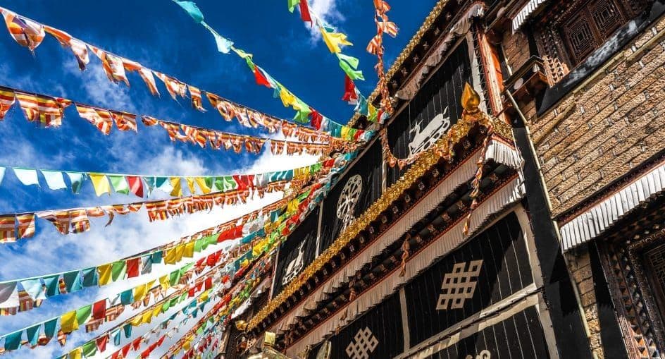 Exterior of a Bhutanese dzong with stone walls, black facade, golden trim, and colorful prayer flags against a blue sky.