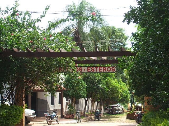 Tropical town entrance with a sign reading Arroyos y Esteros, lush greenery, parked motorcycles, and a small building.
