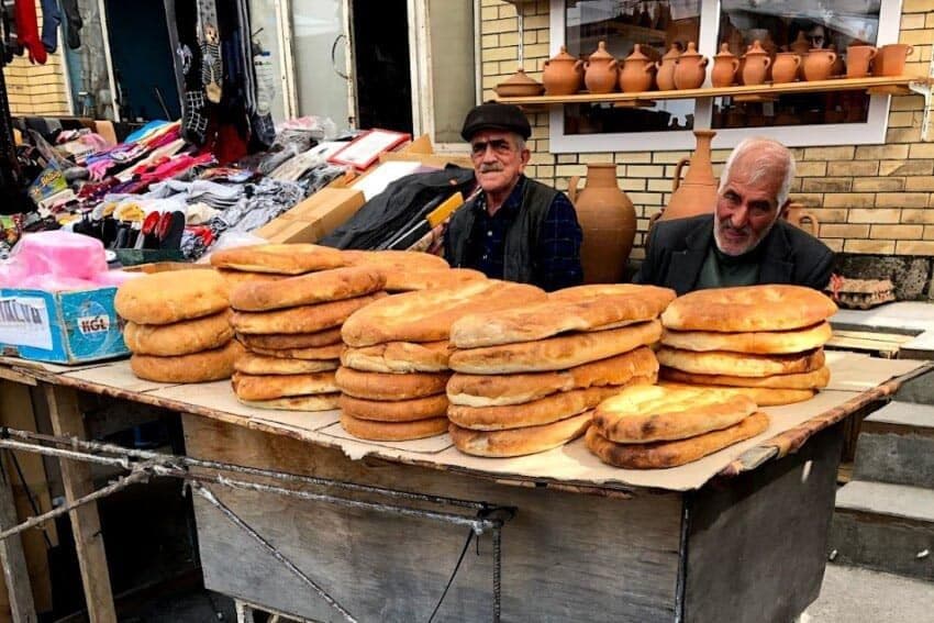 Two vendors sit behind a street table piled with round flatbreads, with pottery and market goods in the background.