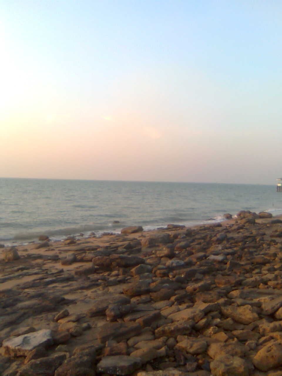 A rocky shoreline meets the calm sea under a soft, pastel sky, capturing the tranquil coastal scenery of Az Zawr beach.