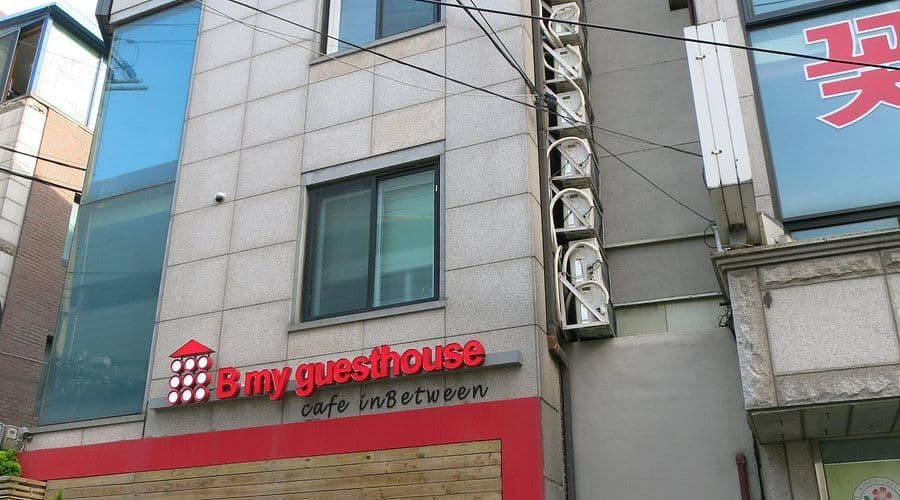 Exterior of a small guesthouse building with a red sign reading 'B my guesthouse cafe inBetween' on a stone facade.