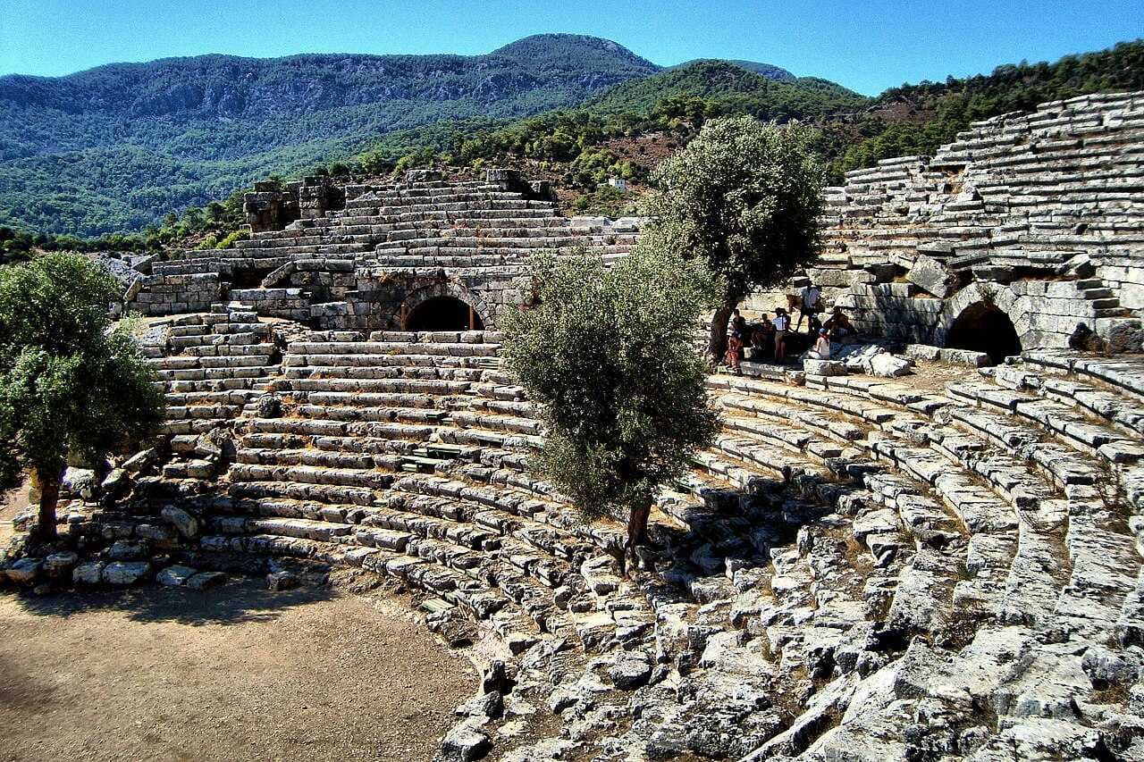 Stone tiered seating of an ancient Roman theatre with olive trees growing in the center, set against a green mountain backdrop.