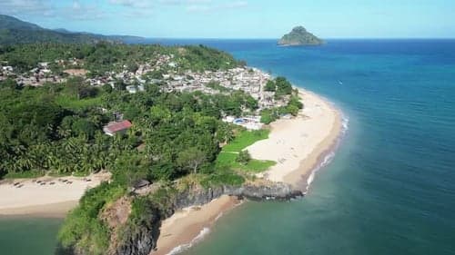 Aerial view of a coastal village on a lush green peninsula with sandy beaches and a distant island in the blue ocean.