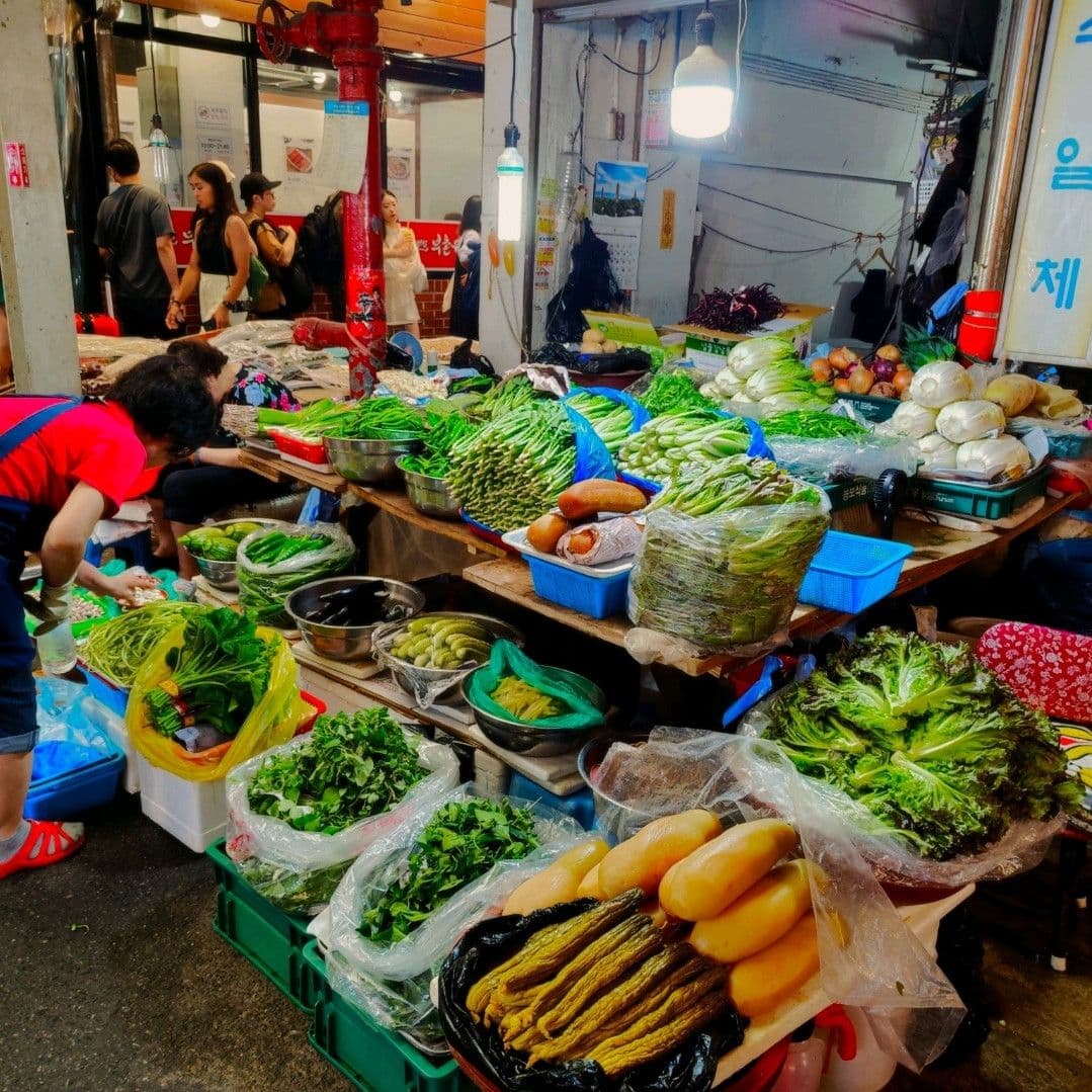 A real photograph of a local market stall displaying fresh vegetables, herbs, and produce, with vendors and shoppers in the background.