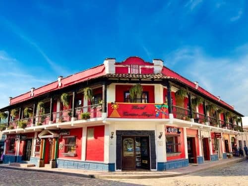 Colorful two-story building with red facade and balconies in a small town plaza under a blue sky.