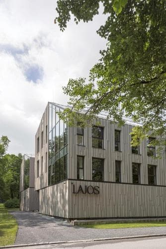 Spring scene at a park entrance in Birštonas, Lithuania showing walking paths, blooming trees, and a contemporary restaurant facade