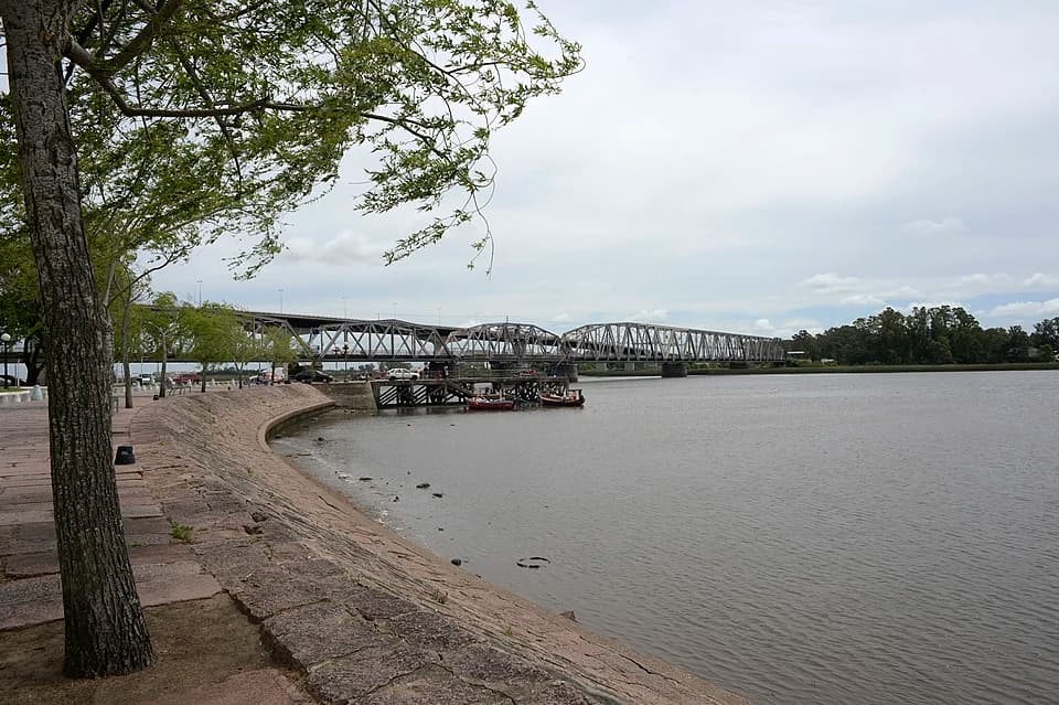 Long steel truss bridge spanning the Santa Lucía River at Santiago Vázquez with stone embankment and trees in the foreground