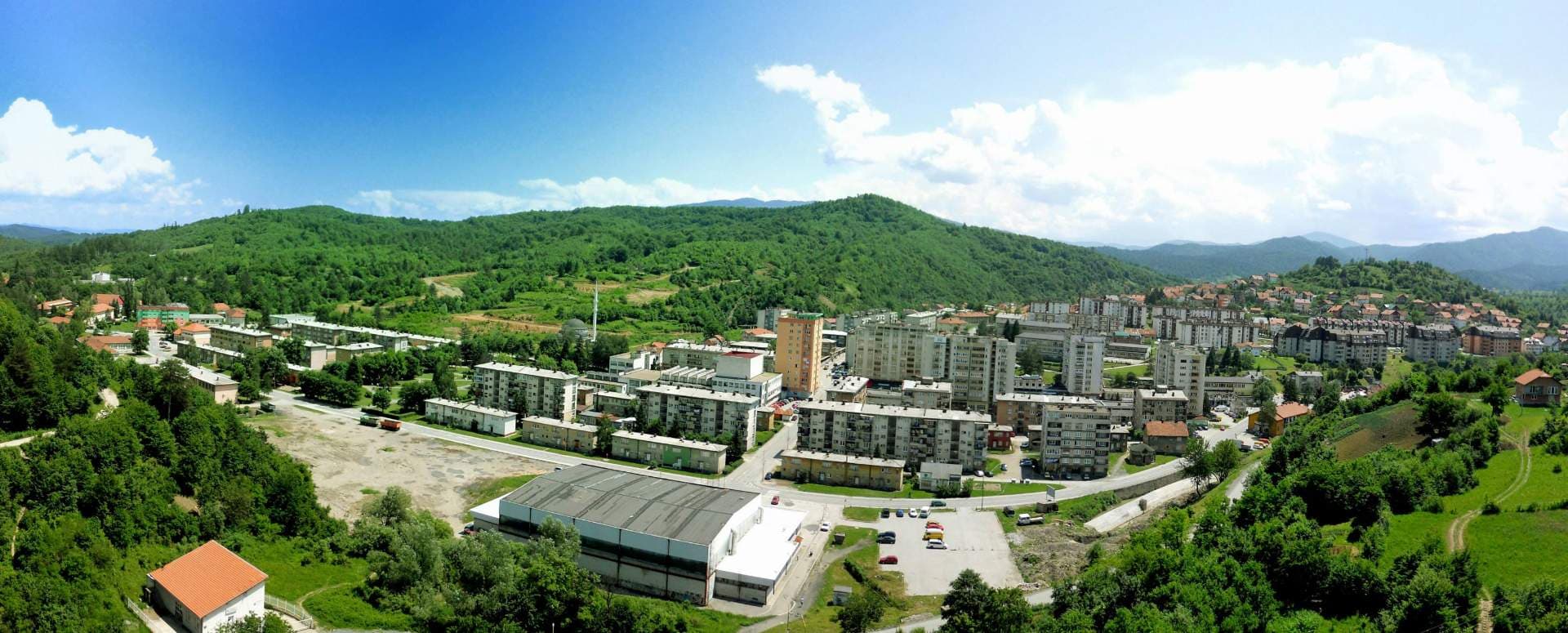 Panoramic aerial view of a town nestled in a green valley with forested hills, featuring apartment blocks and industrial buildings under a blue sky.
