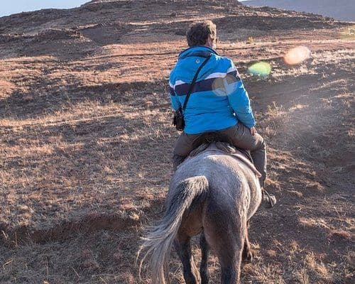 A person in a blue jacket rides a horse across dry, grassy highland terrain with rolling hills in the background.