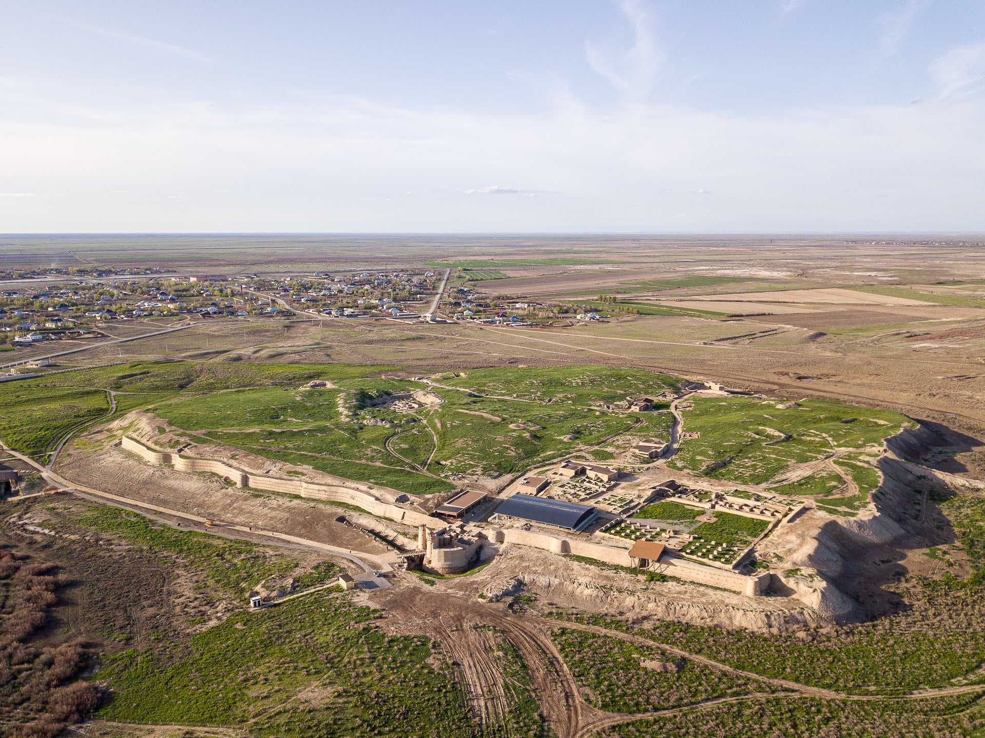 Aerial view of ancient stone settlement ruins with walls and foundations on a grassy steppe, near a small town and open fields.