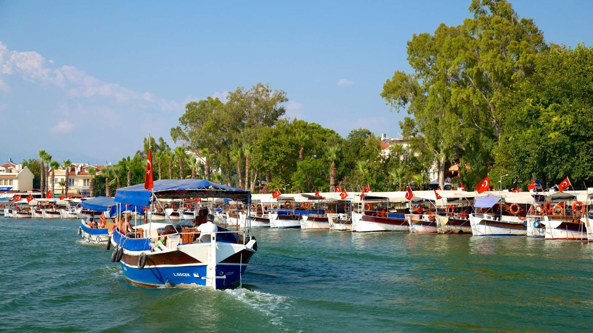 Tour boats with Turkish flags navigate a river channel near a waterfront town with palm trees and buildings.