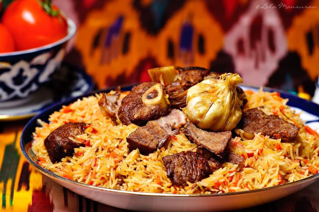 A close-up shot of a traditional Tajik plov dish served on a platter, featuring yellow rice, chunks of meat, and a whole roasted garlic bulb.