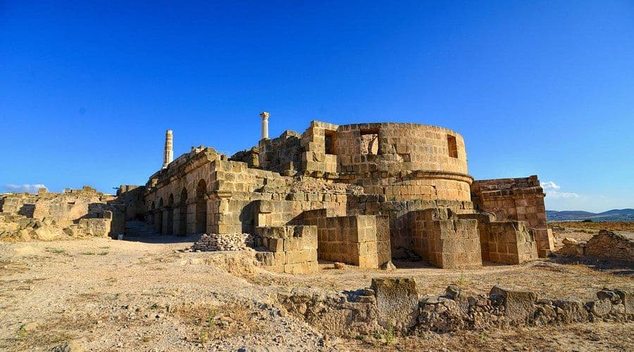 Sun-bleached Roman ruins with a circular structure and columns at the Uthina (Oudhna) site in Ben Arous under a clear blue sky.
