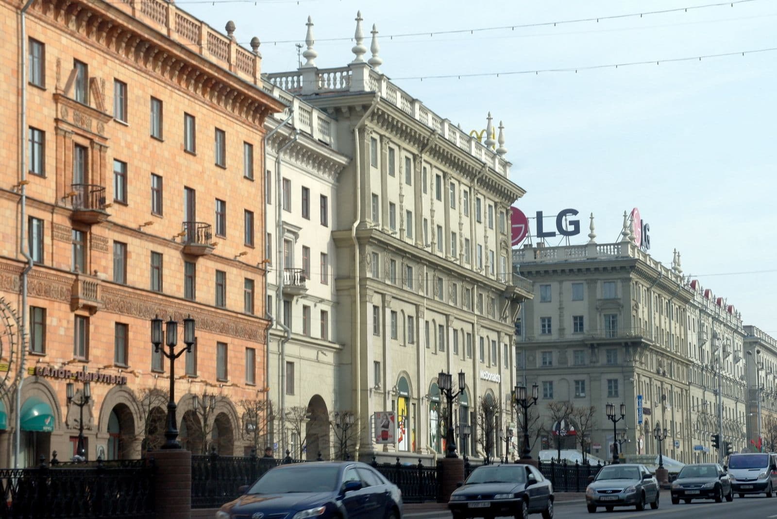 Wide streetscape of ornate multi-story buildings with arched ground floors, street lamps, and cars on a city road.