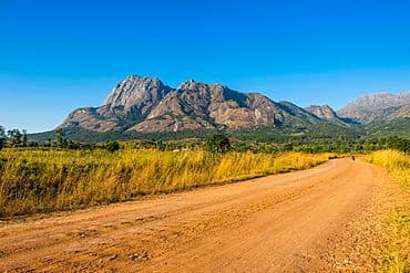 A wide landscape view of Mount Mulanje granite mountains near Phalombe, featuring a dirt road winding through golden grasslands under a clear blue sky.