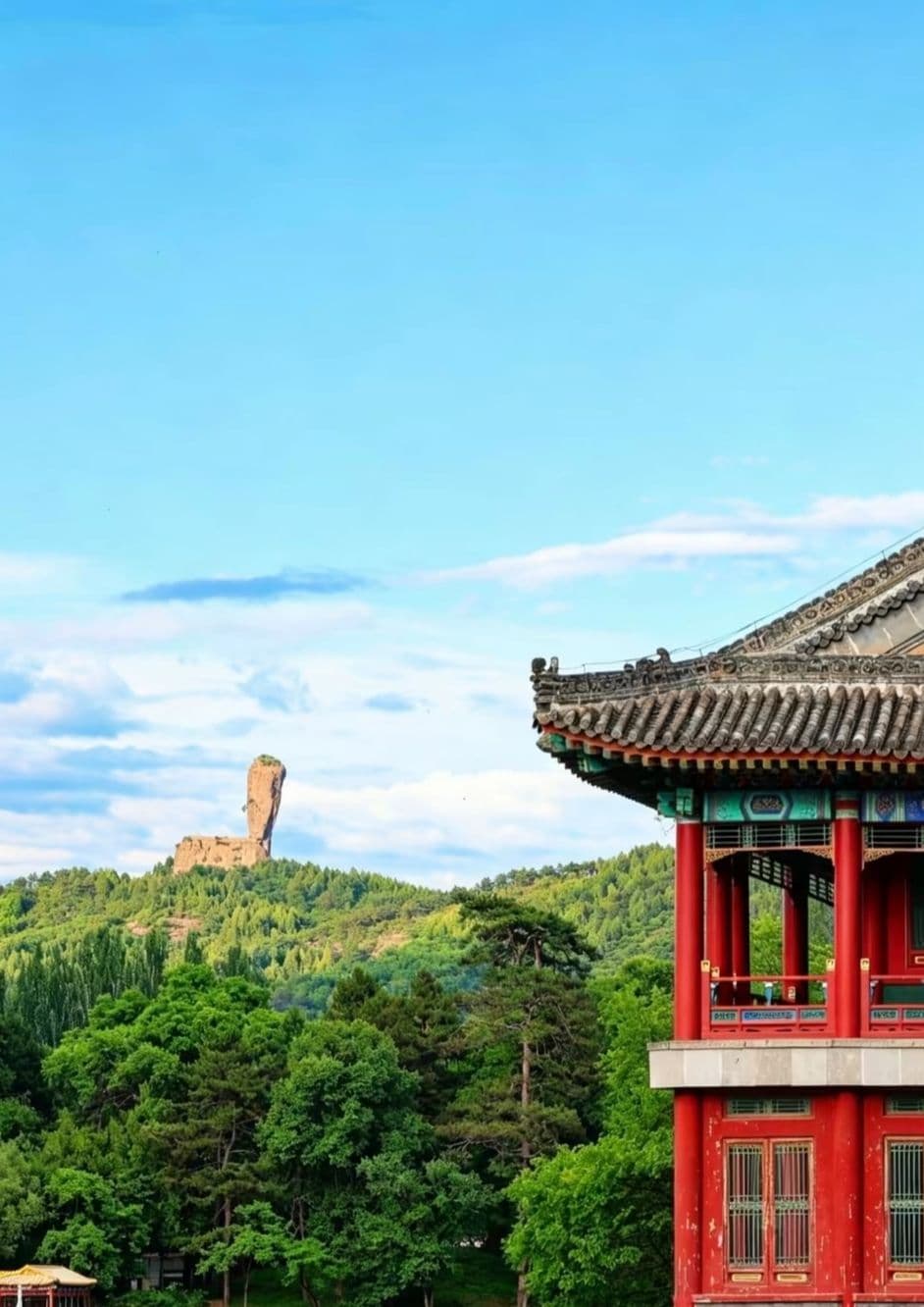 Traditional red pavilion with tiled roof in foreground, forested hills and a distinctive rock formation under blue sky.