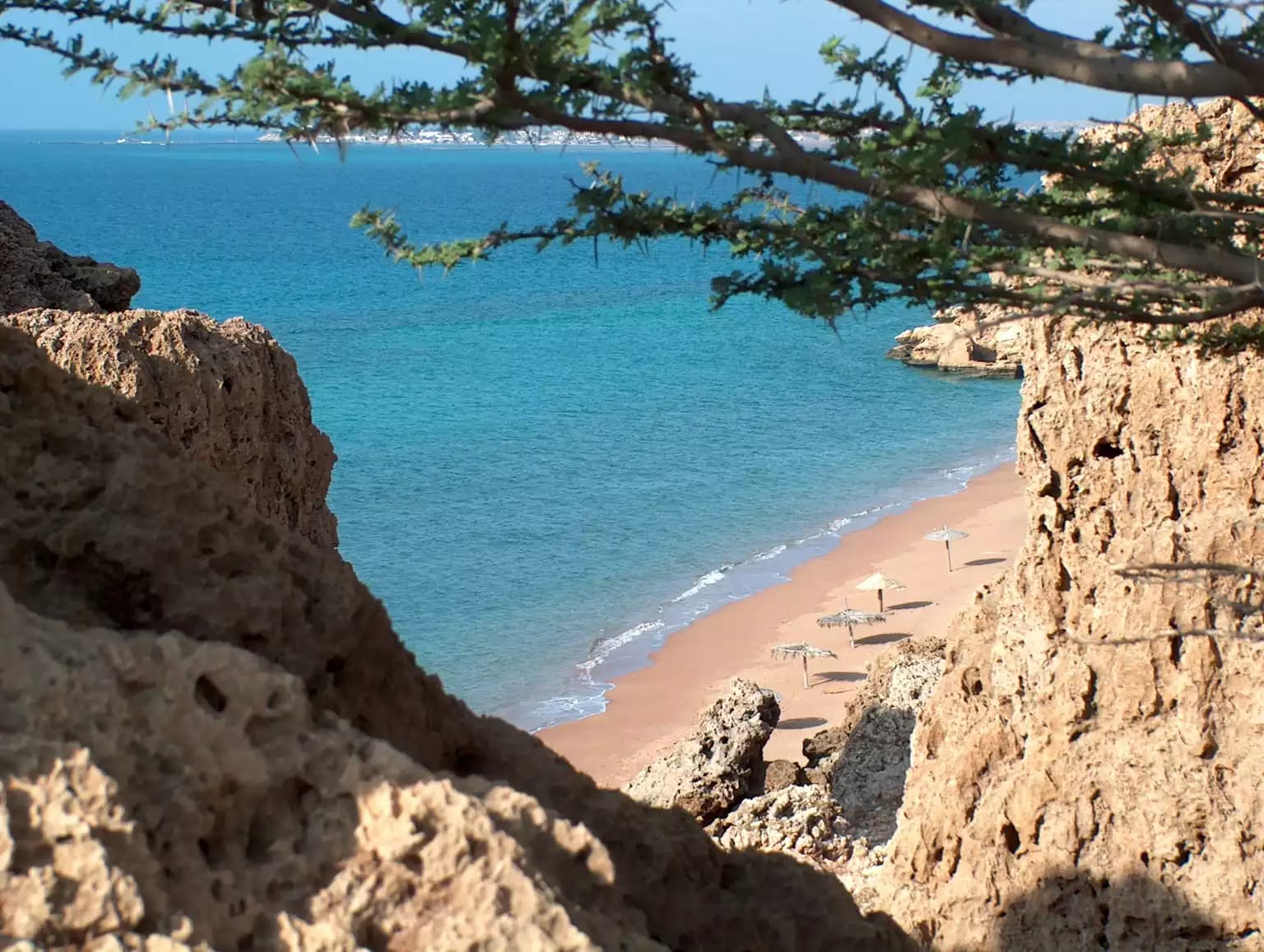 A scenic view of a secluded sandy beach with thatched umbrellas, framed by rocky cliffs and tree branches, overlooking turquoise waters.