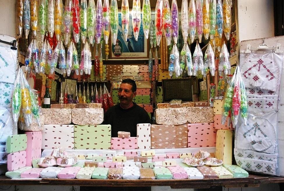 A man stands behind a market stall displaying colorful packaged soaps, hanging candy, and embroidered textiles in a Mauritanian souk