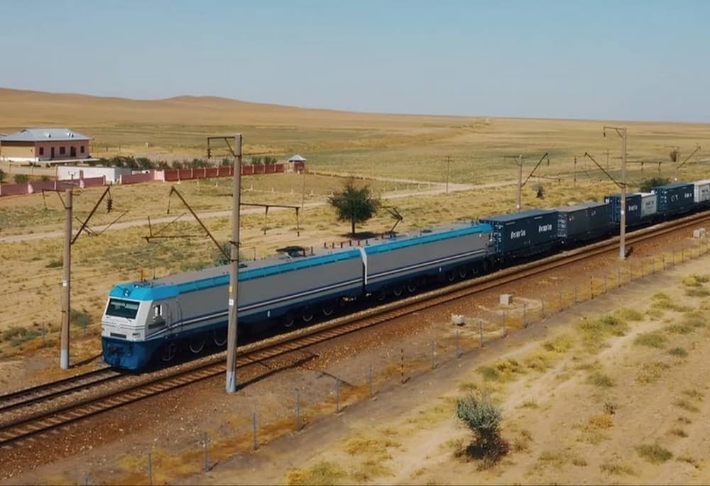 A blue and grey electric locomotive hauling freight containers travels along railway tracks through the arid, golden landscape of Turkmenistan.