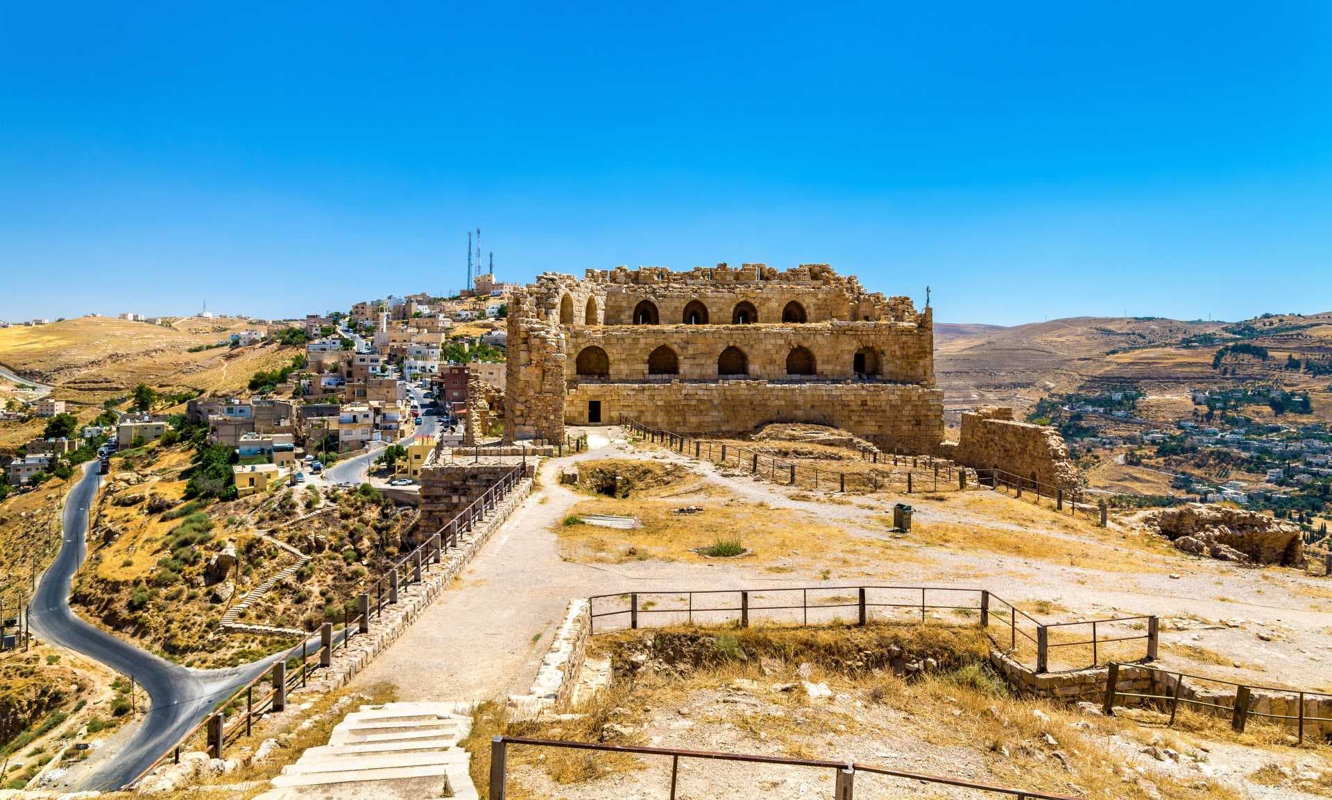 Sunlit stone ruins with arched windows overlook a village in the Karak highlands, set against a clear blue sky and dry, hilly terrain.