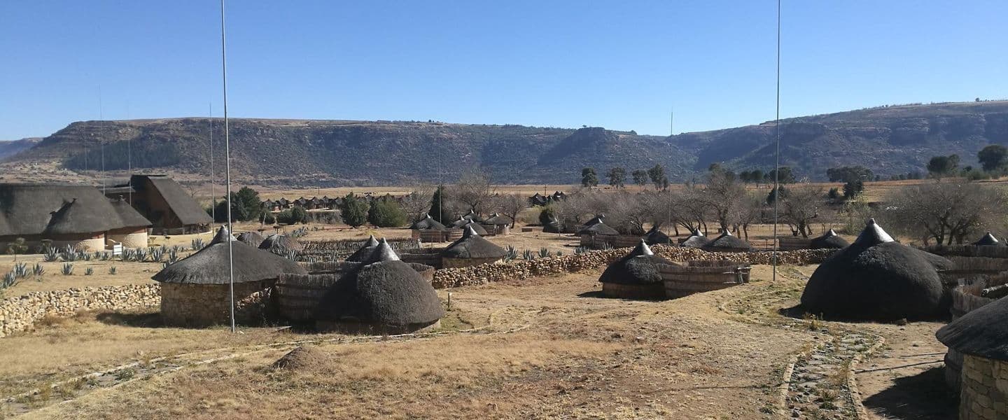 Wide high-altitude grassland vista with traditional thatched huts, dry autumn light, and mountain slopes in the background.