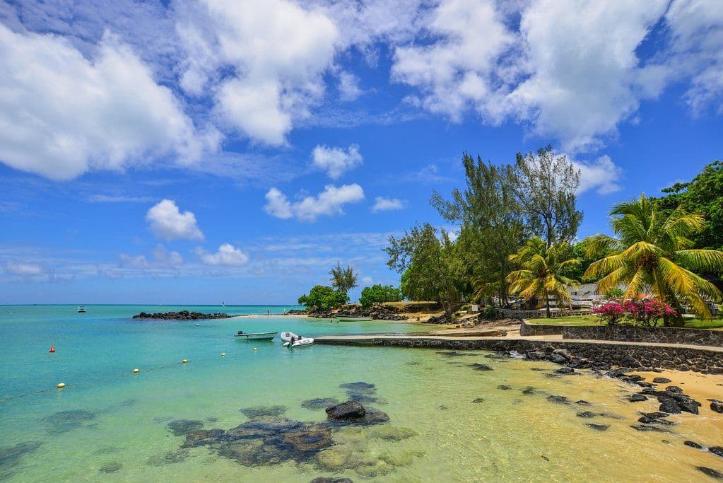 Coastal lagoon with turquoise water, small boats, rocky shoreline, palm trees, and flowering plants under a blue sky.