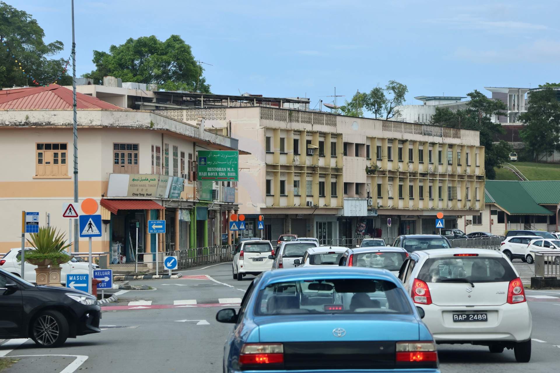 Busy street scene in Tutong town featuring traffic, traditional shophouses, and modern buildings under a clear sky.