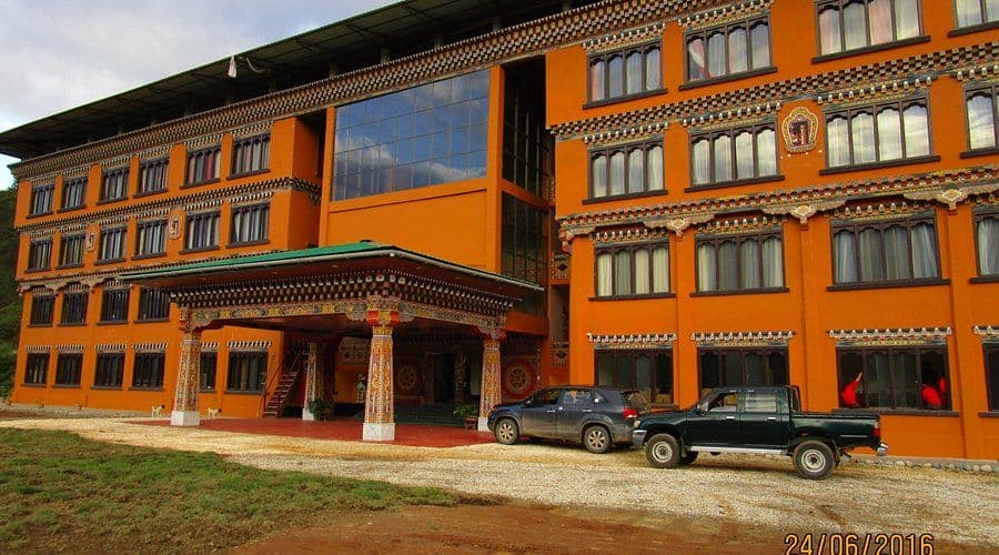 Exterior view of a large orange building featuring traditional Bhutanese architectural details, with vehicles parked on a gravel driveway.