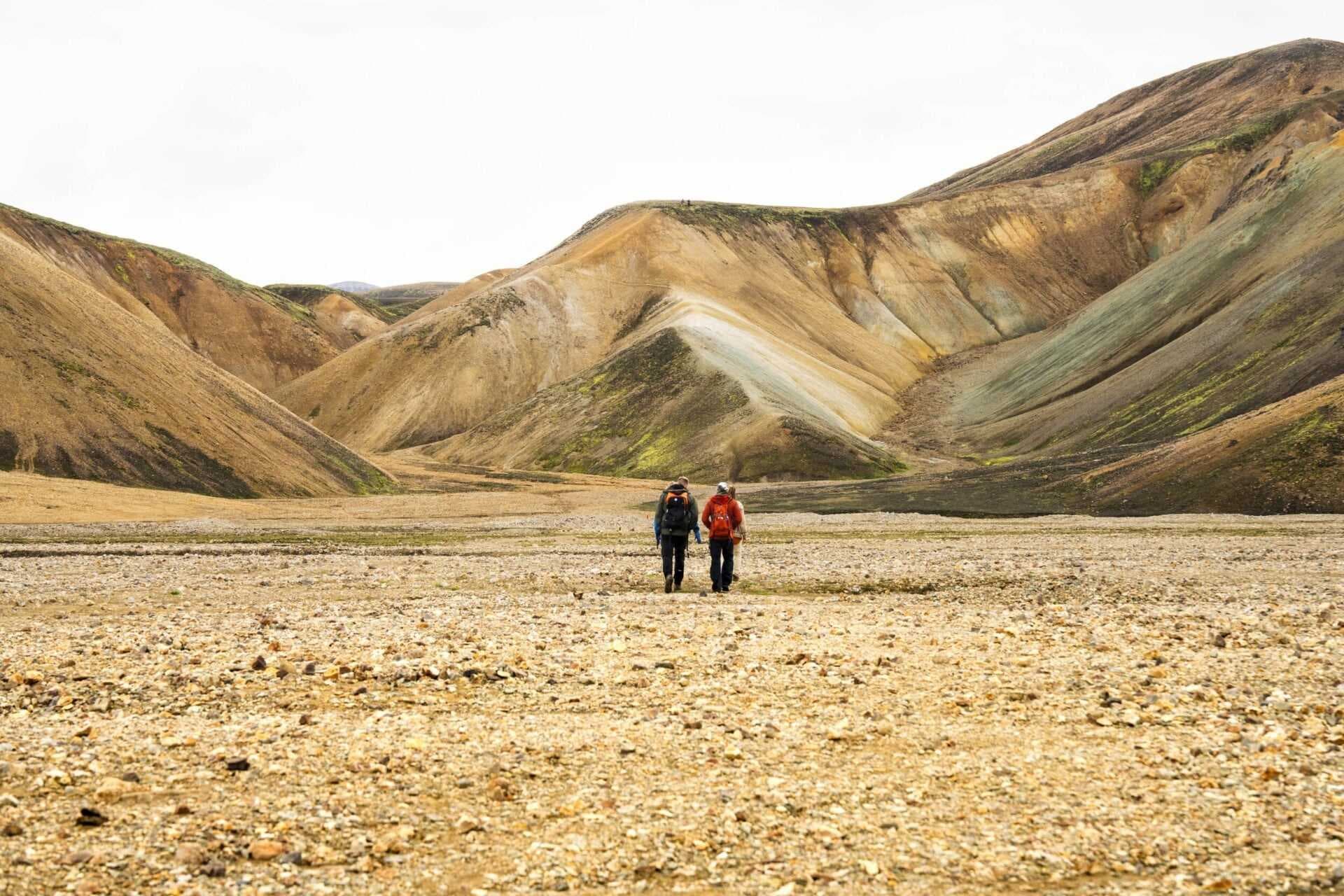 Two hikers with backpacks walk across a rocky highland plain toward multicolored hills under an overcast sky.