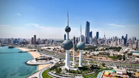 Wide aerial view of Kuwait City skyline near Ash Shāmīyah featuring the Kuwait Towers, waterfront, and modern high-rise buildings under a clear blue sky