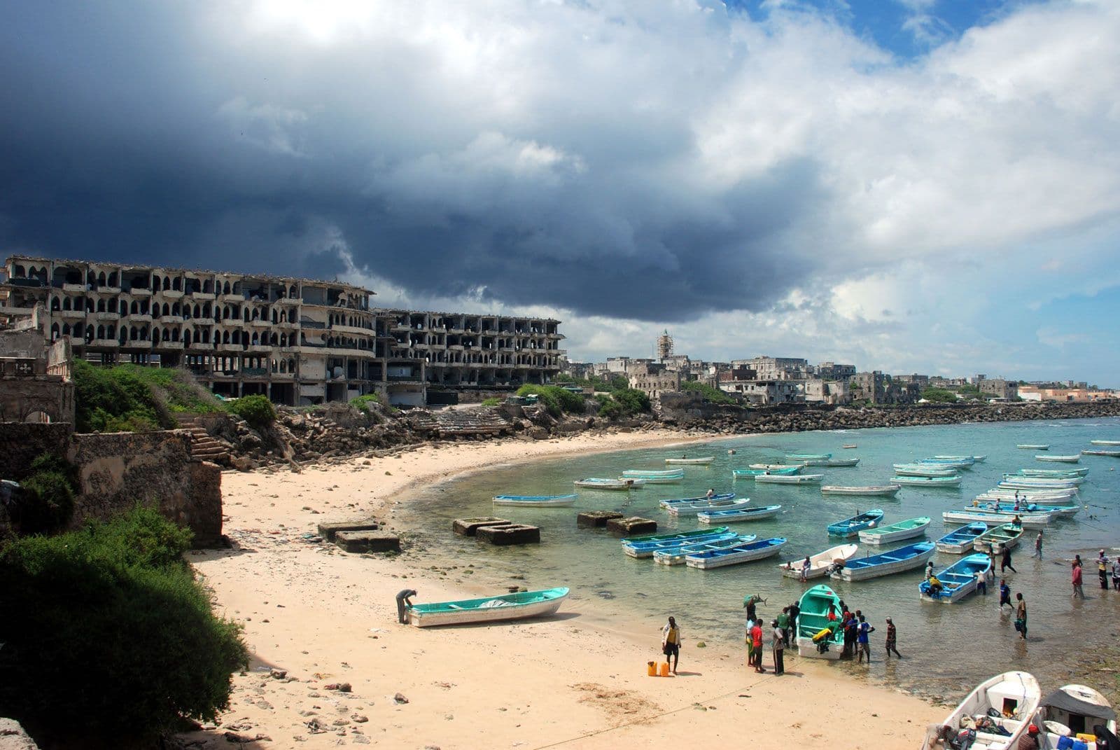 Coastal shoreline with sandy beach, numerous small boats in calm water, ruined multi-story buildings, and scattered people near boats under dramatic storm cloud