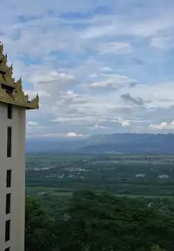 A view from a high vantage point showing a golden architectural spire on the left, overlooking a vast green valley and distant mountains under a cloudy sky.