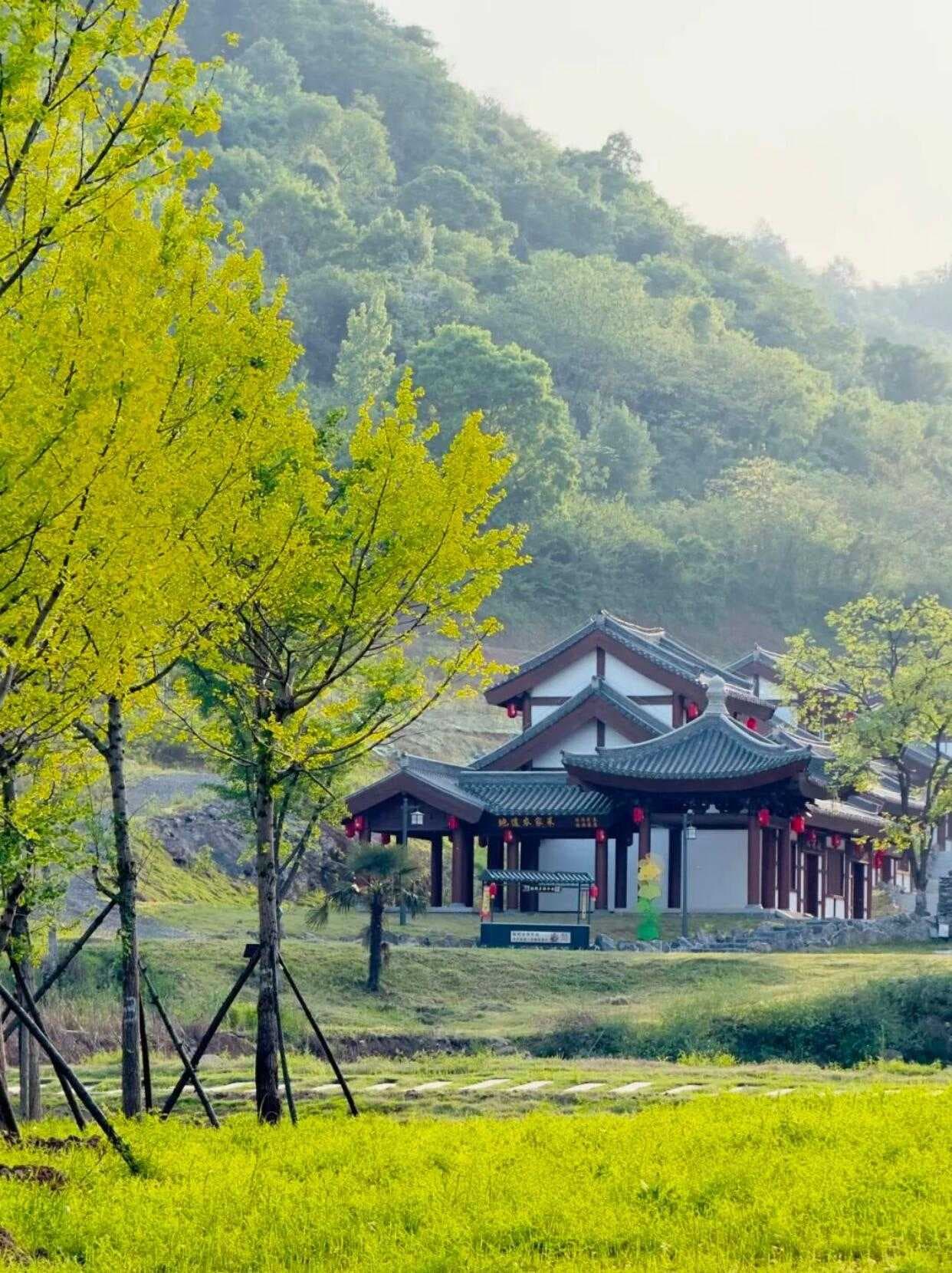 Traditional East Asian pavilion with red lanterns set in a green field, backed by forested hills and young trees.