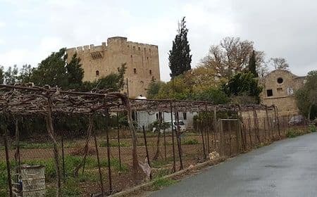 A stone castle and rural buildings stand behind a vineyard trellis in a hilly countryside setting near Soúni-Zanakiá.