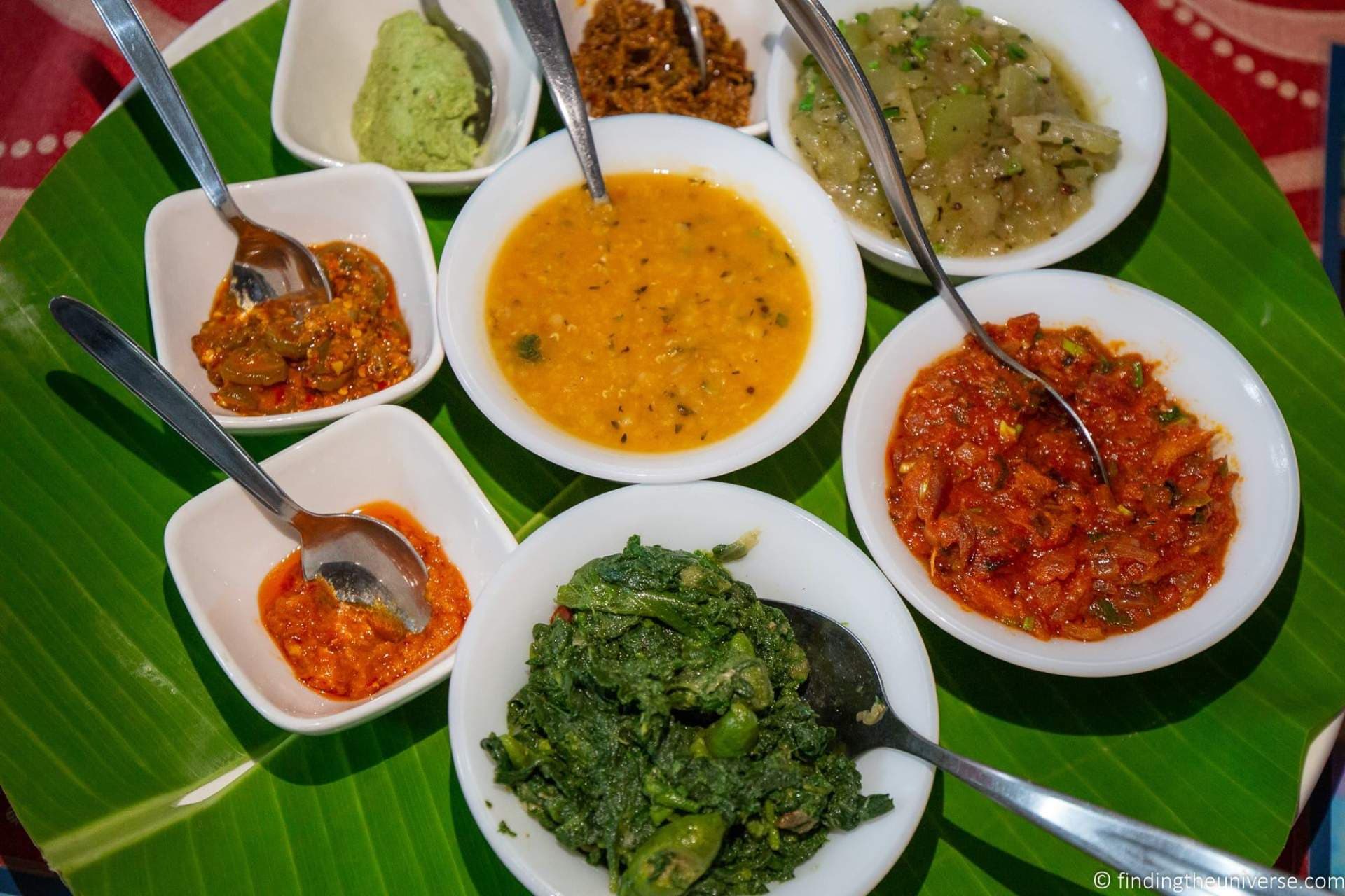 Multiple small white bowls of colorful curries and chutneys arranged on a green banana leaf with spoons.